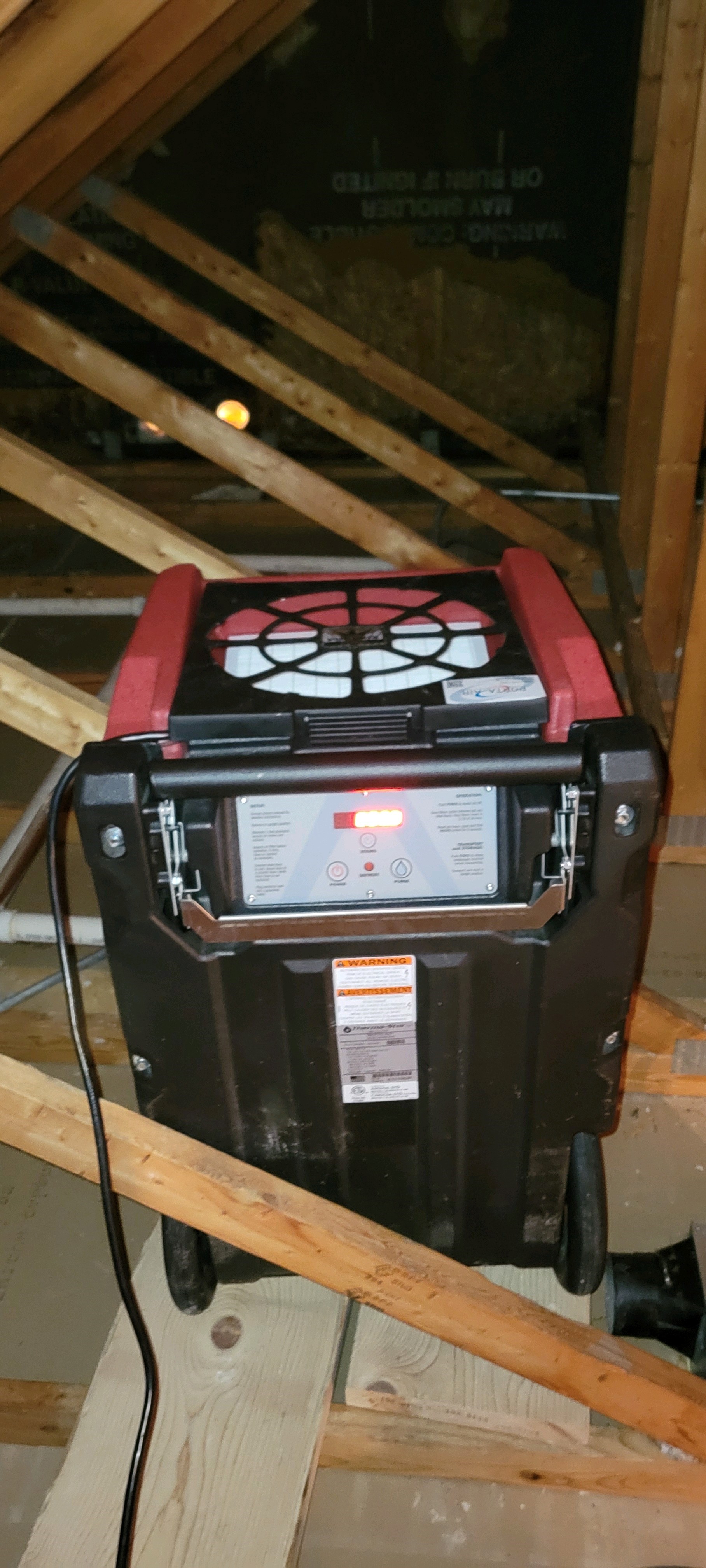 A dehumidifier positioned on the joists in an attic below the roof rafters removing humidity from the attic. 