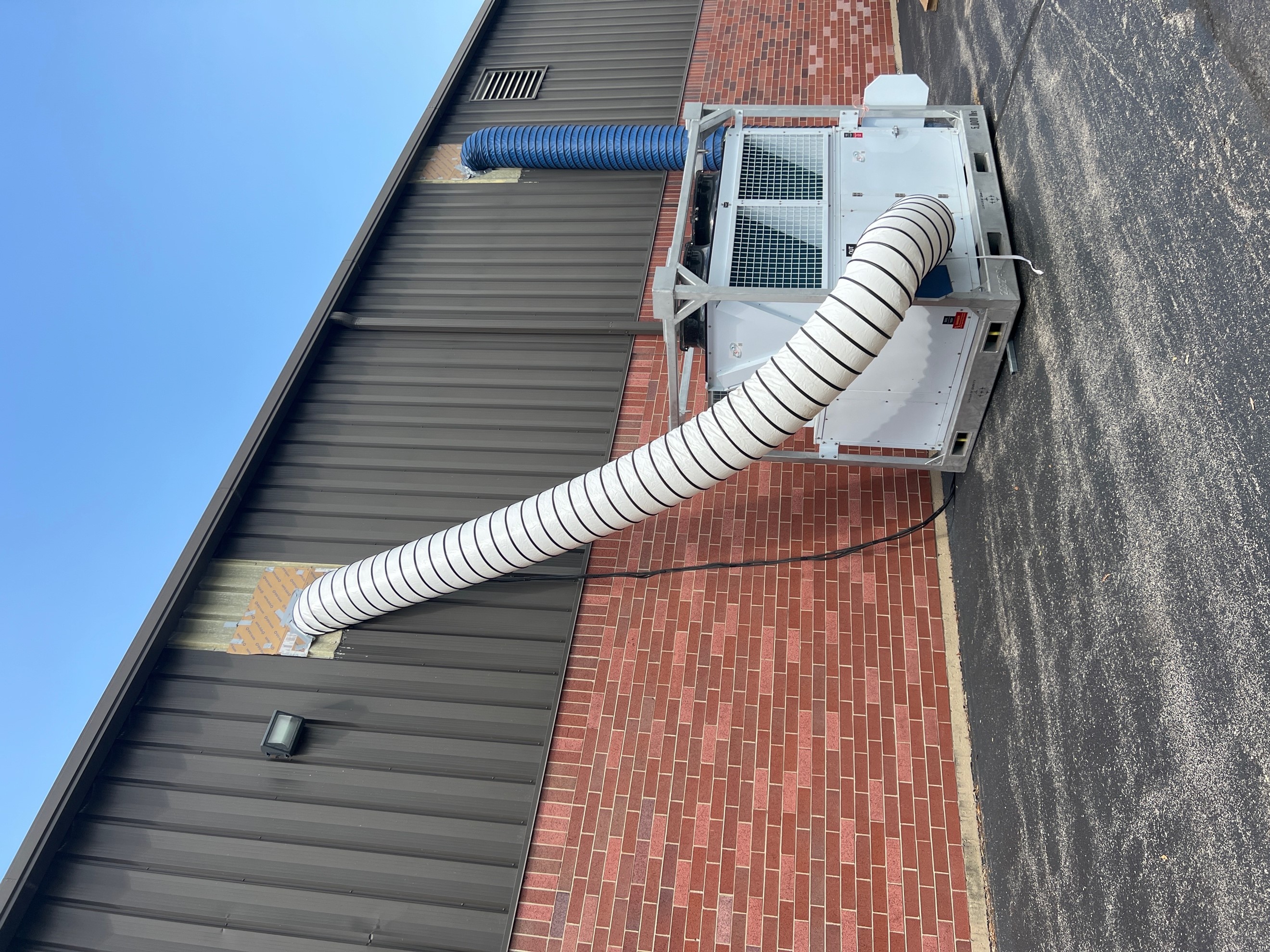 A skid mounted mobile air conditioner positioned in a parking lot ducted to two upper building openings using white and blue ductwork. 