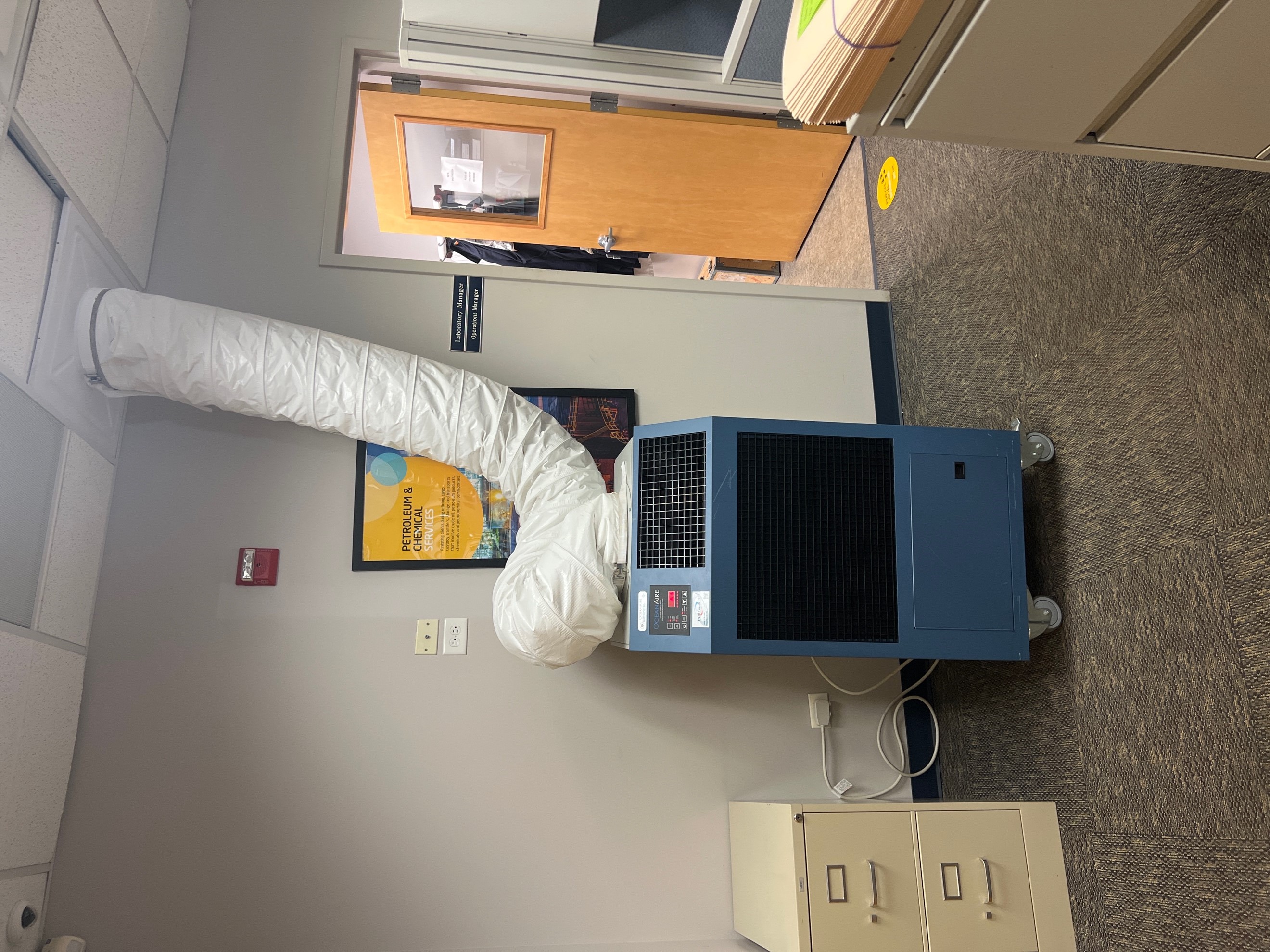 A Portable Heat Pump Rental plugged into a wall outlet, spot cooling an office space using white flexible duct directed to a drop ceiling. 